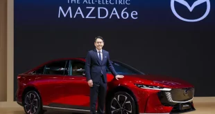 The All-Electric Mazda6e สีแดงจัดแสดงบนเวที พร้อมบุคคลยืนข้างรถและโลโก้ Mazda