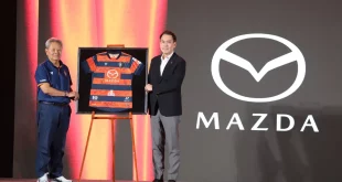 ผู้ร่วมงานยืนข้างเสื้อแข่งขันสโมสรนครราชสีมา มาสด้า เอฟซี ที่มีโลโก้ Mazda บนเวที