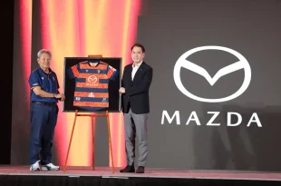 ผู้ร่วมงานยืนข้างเสื้อแข่งขันสโมสรนครราชสีมา มาสด้า เอฟซี ที่มีโลโก้ Mazda บนเวที