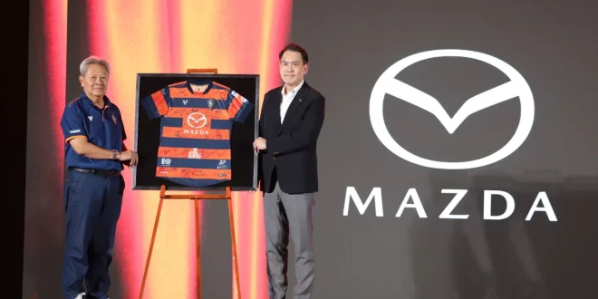 ผู้ร่วมงานยืนข้างเสื้อแข่งขันสโมสรนครราชสีมา มาสด้า เอฟซี ที่มีโลโก้ Mazda บนเวที