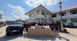 กลุ่มผู้ร่วมส่งมอบสิ่งของช่วยเหลือน้ำท่วมยืนด้านหลังกล่องสิ่งของ น้ำดื่ม และถุงอีซูซุ โดยมีรถอีซูซุจอดด้านข้าง