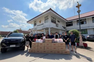 กลุ่มผู้ร่วมส่งมอบสิ่งของช่วยเหลือน้ำท่วมยืนด้านหลังกล่องสิ่งของ น้ำดื่ม และถุงอีซูซุ โดยมีรถอีซูซุจอดด้านข้าง