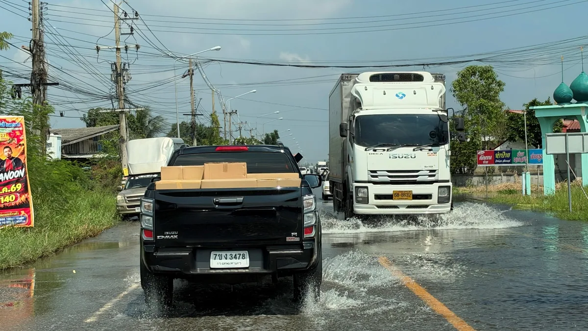 รถอีซูซุบนถนนน้ำท่วมพร้อมสิ่งของบรรทุก รถกระบะอีซูซุสีดำบรรทุกกล่องสิ่งของขับบนถนนที่มีน้ำท่วมขัง พร้อมรถบรรทุกอีซูซุสีขาววิ่งสวนมา