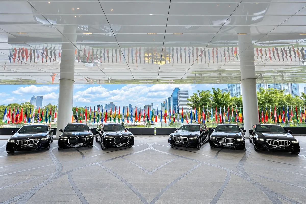 รถยนต์ BMW จอดเรียงกันสำหรับข่าวความร่วมมือ MGC-ASIA รถยนต์ BMW สีเข้ม 6 คันจอดเรียงกันใต้หลังคาอาคาร โดยมีธงนานาชาติอยู่ด้านหลัง