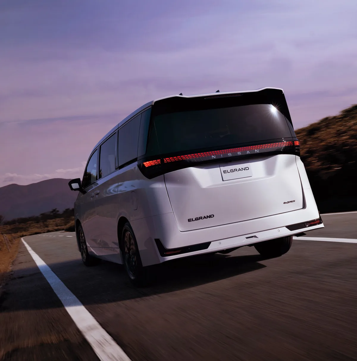 Nissan Elgrand มุมหลังขณะวิ่งบนถนน Nissan Elgrand สีขาวมุมด้านหลังขณะวิ่งบนถนน เห็นไฟท้ายแนวยาว ป้ายชื่อ Elgrand และสัญลักษณ์ e-4ORCE