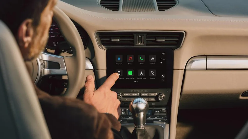 หน้าจอเมนู Porsche PCCM Plus ผู้ขับขี่แตะหน้าจอ PCCM Plus ในห้องโดยสาร Porsche ซึ่งแสดงเมนูและไอคอน Apple CarPlay กับ Android Auto