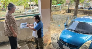 เจ้าหน้าที่ซูซูกิอธิบายเอกสารให้ผู้หญิงข้างรถ Suzuki สีน้ำเงินที่จอดอยู่ริมอาคาร